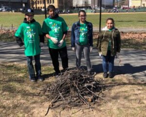 Four students in green t-shirts smile behind of a pile of brush, cleaned up from the local park.